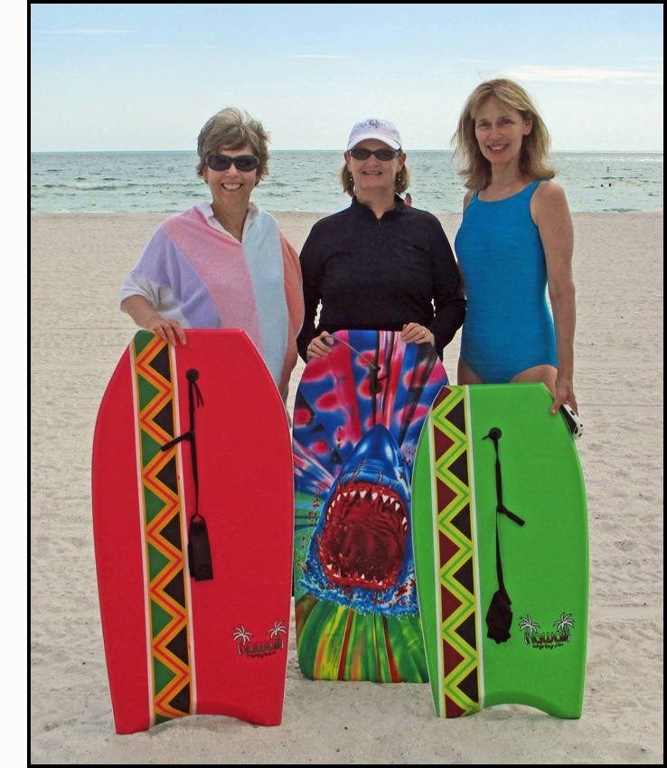 Photo shows 3 women smiling broadly and standing on the beach behind their boogy boards.  Dona is on the left wearing sunglasses and a white, pink and purple terrycloth coverall, her boogy board is red with a stripe of green, black, orange and red zig-zags.  Liz is in the middle wearting a white baseball cap and sunglasses with a black long-sleeved shirt, her boogie board has a large shark's open jaw and bright colors.  Marjie is on the right wearing a blue bathing suit, her boogie board is bright green with a stripe of green, black, green and brown zig-zags. The ocean behind them has no waves higher than about a foot.
